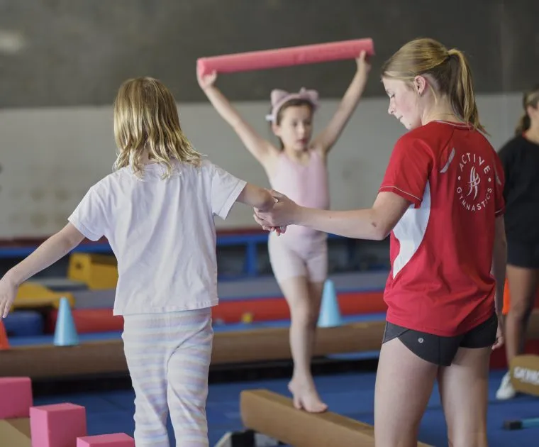 Coach holding a gymnast's hand as they balance across a beam stepping over foam blocks.