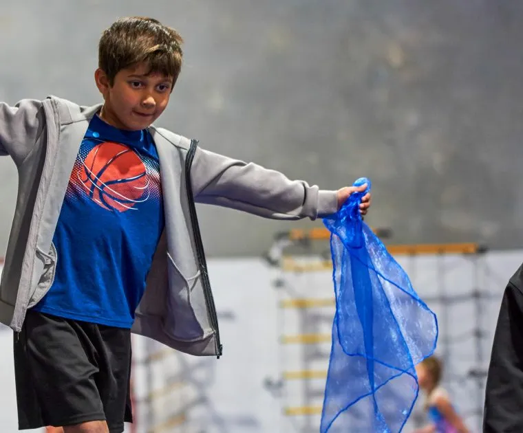 A gymnast walks across a beam holding two coloured scarfs. A coach is standing next to them smiling and helping them.