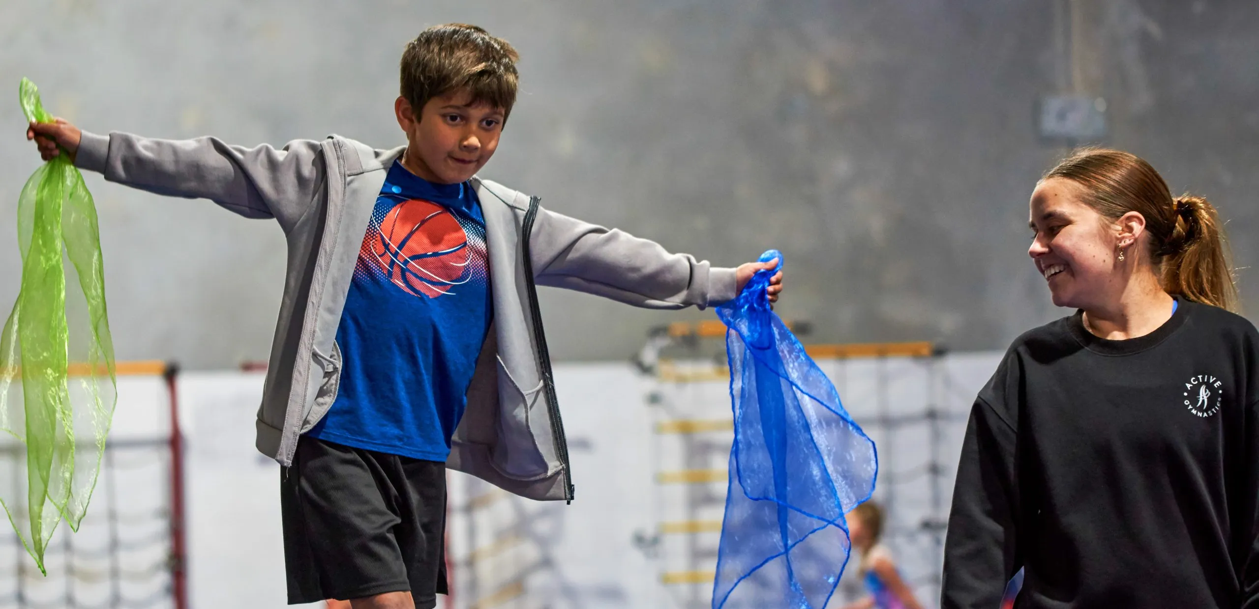 A gymnast walks across a beam holding two coloured scarfs. A coach is standing next to them smiling and helping them.